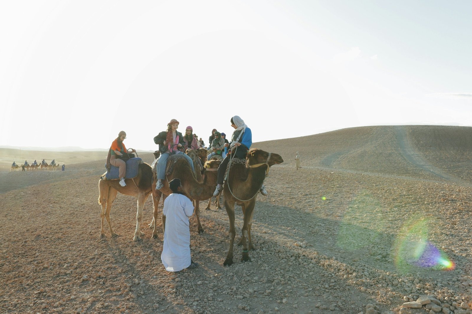 Tourists riding camels in Agafay Desert guided by locals in traditional Moroccan clothing under soft desert sunlight.