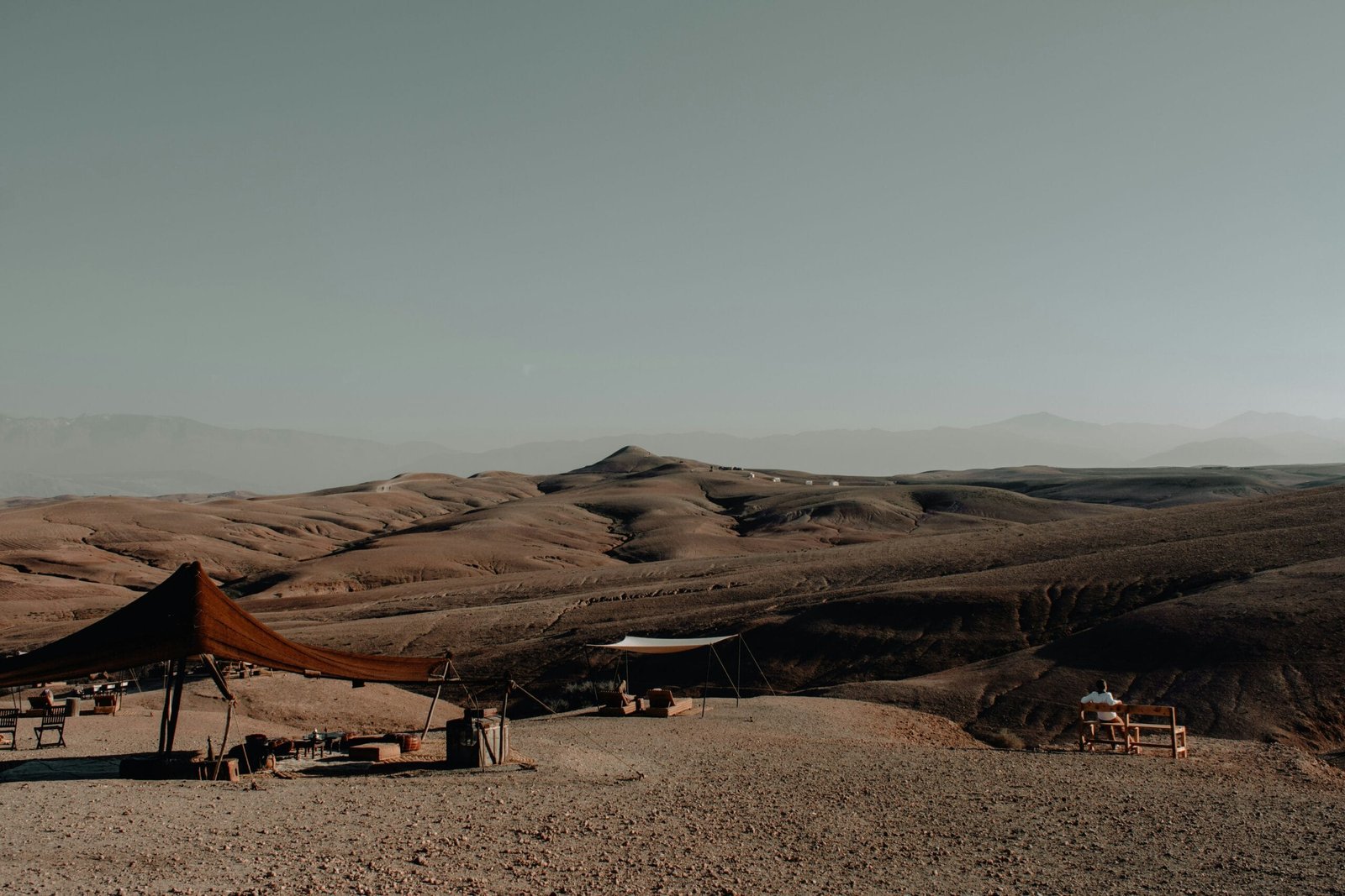 Scenic view of Agafay Desert’s rocky hills with luxury desert camp tents and seating areas, with Atlas Mountains visible in the distance.